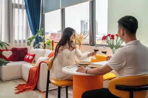 Una pareja cenando en una mesa con vistas a la ciudad, un sofá con almohadas en forma de corazón y una planta en el alféizar de la ventana.