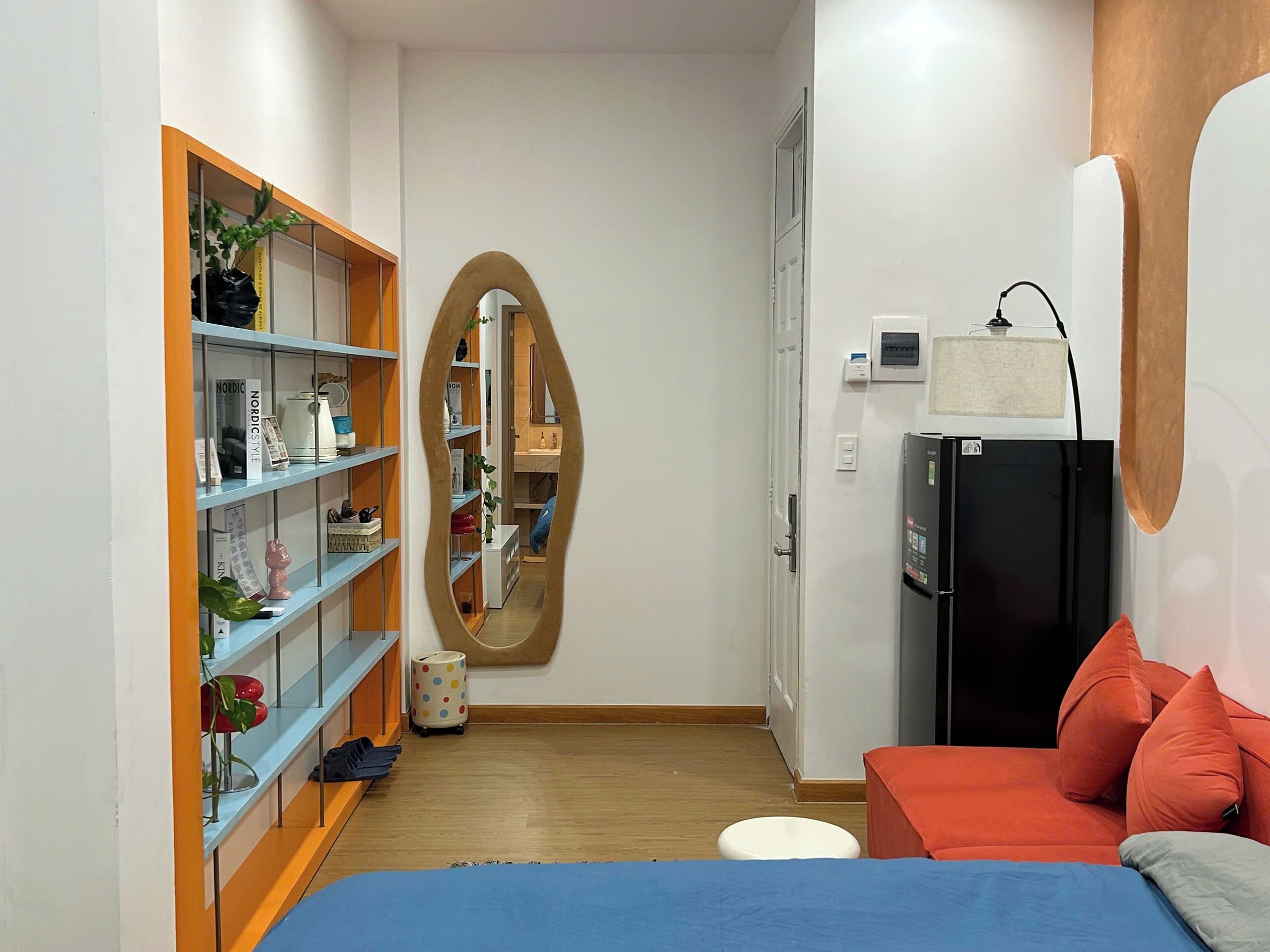A compact room with a bed, a bookshelf, a mirror, and a refrigerator.