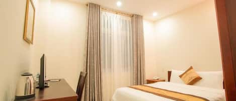 A hotel room with a bed, a desk with a kettle, a chair, a television, and a curtain.