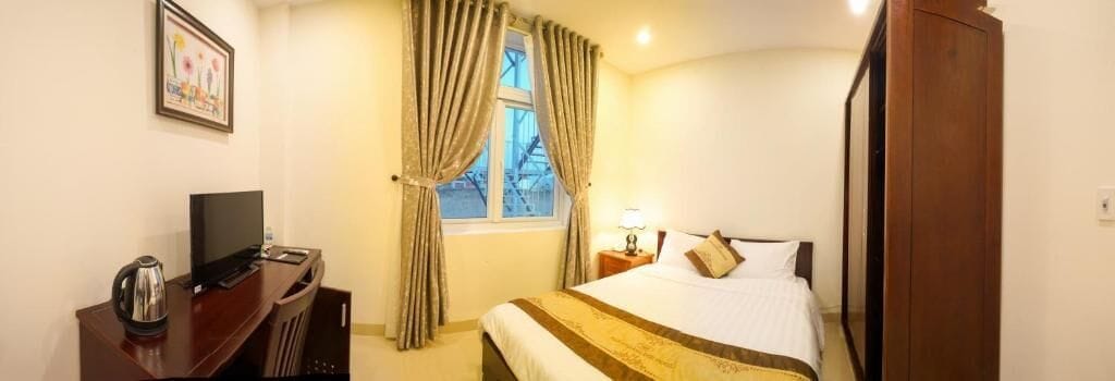 Habitación de hotel con cama, escritorio con televisor, ventana con cortinas y mesita de noche con lámpara.