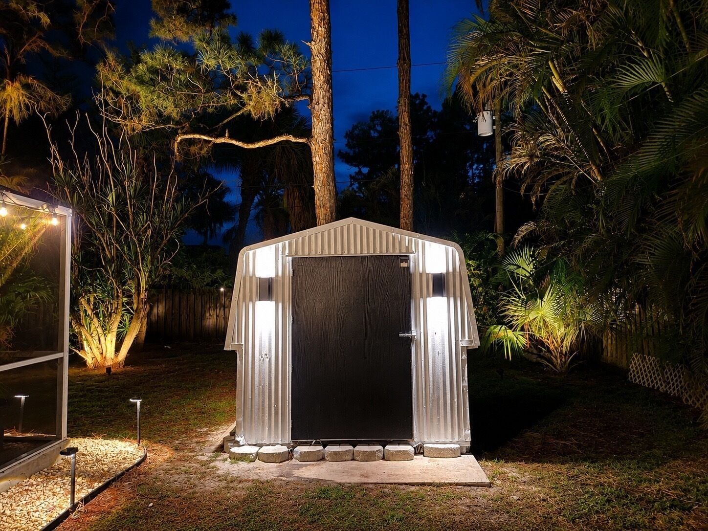 A metal shed with a dark door, surrounded by a landscaped garden with palm trees and a wooden fence.