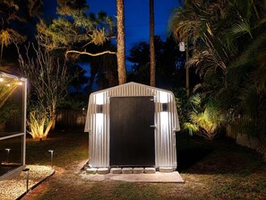 A metal shed with a dark door, surrounded by a landscaped garden with palm trees and a wooden fence. - Palm Vibe House - Pool • Near Beach • Florida (Port St. Lucie)