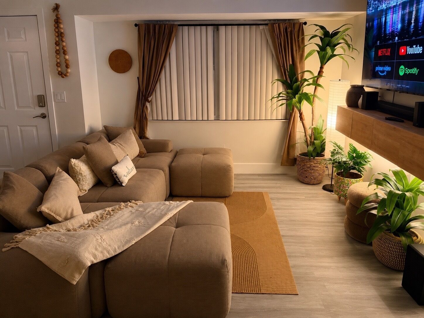 A living room with a sectional sofa, a flat-screen TV, and potted plants.