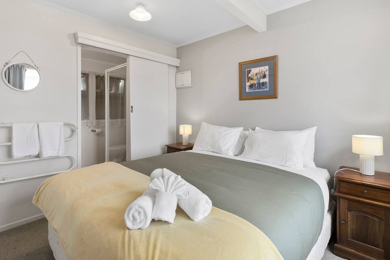 A hotel room with a bed, a bedside table with a lamp, a bathroom with a shower, and a mirror on the wall.