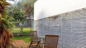 Eine Terrassenfläche mit zwei Holzstühlen und einem Tisch, einer weißen Wand und einem Garten mit Farne und einem Baum.