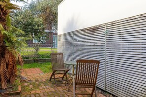 Eine Terrassenfläche mit zwei Holzstühlen und einem Tisch, einer weißen Wand und einem Garten mit Farne und einem Baum.