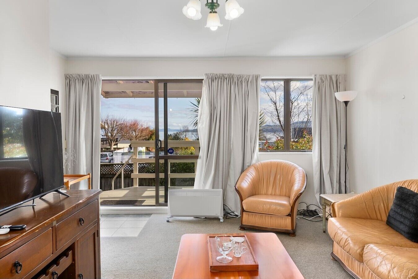 A modern living room with a television, leather sofa, and a dining area visible through large windows.