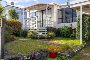 Una casa moderna con un porche cubierto, un pequeño jardín con diversas plantas y una maceta roja en primer plano.