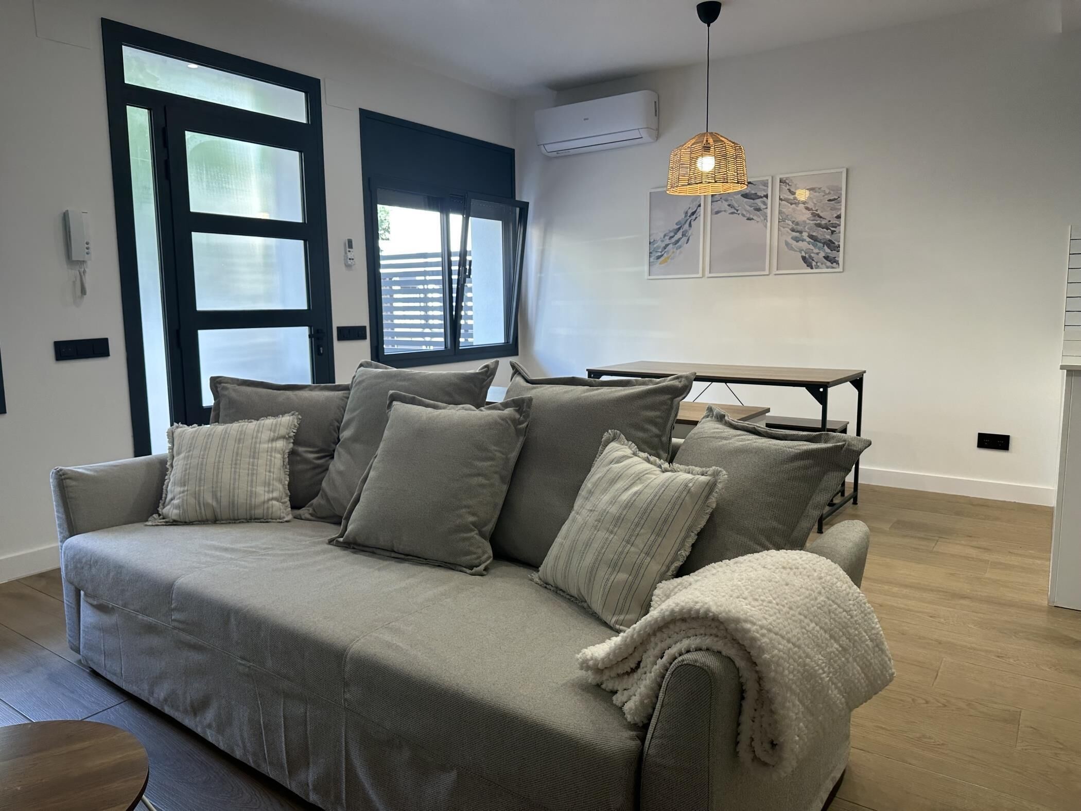 A modern living room with a grey sofa, a wooden coffee table, and a dining table with chairs. There is a large window with blinds, an air conditioning unit, and a hanging pendant light.