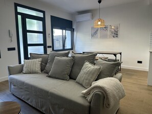 A modern living room with a grey sofa, a wooden coffee table, and a dining table with chairs. There is a large window with blinds, an air conditioning unit, and a hanging pendant light. - Ground floor with A/C and terrace only 250m beach (Calella)