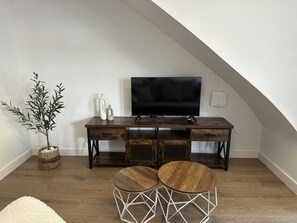 A modern loft bedroom with a sloped ceiling, a wooden desk with a flat-screen TV, a potted plant, and two round wooden tables with geometric legs. - Ground floor with A/C and terrace only 250m beach (Calella)