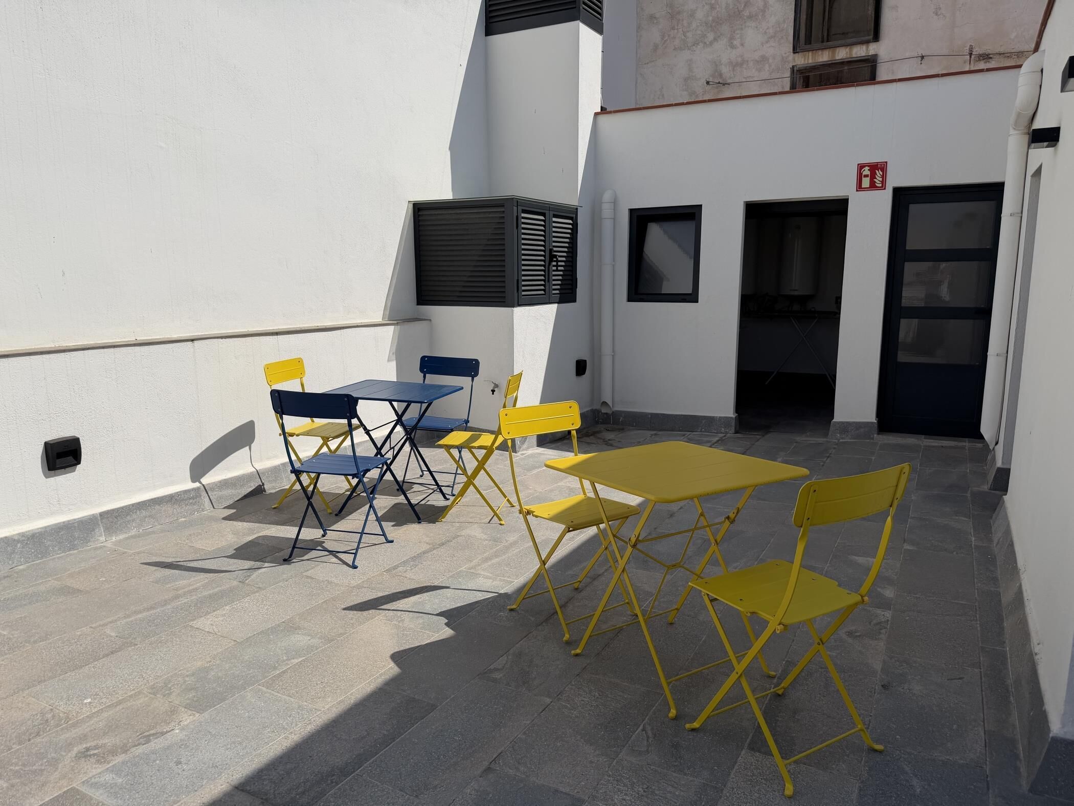 Un espace extérieur aménagé avec des chaises et des tables jaunes et bleues, un panneau indiquant la sortie de secours et l’entrée du bâtiment.