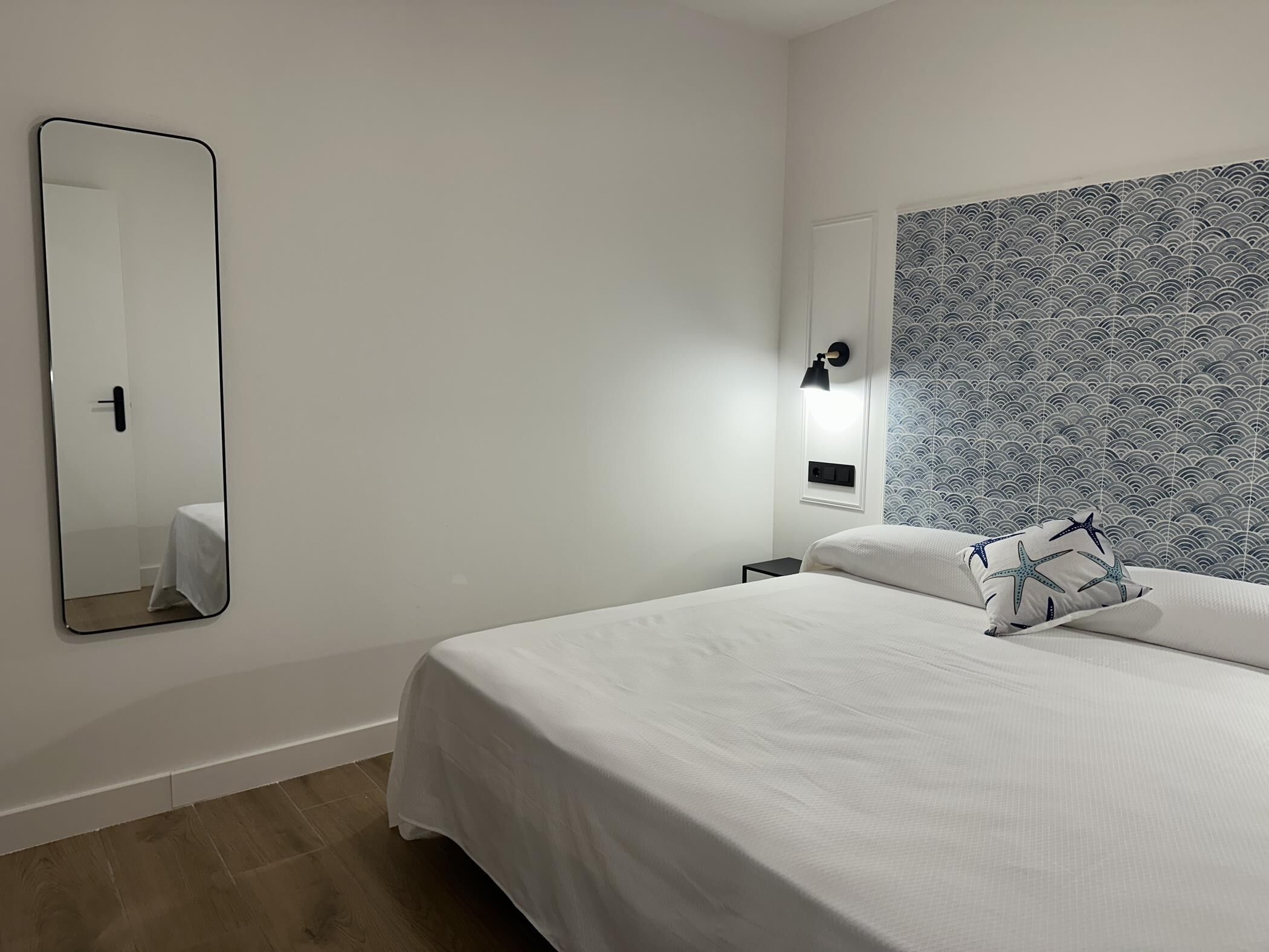 A hotel room with a large bed, a bedside table, a nightstand, a wall-mounted mirror, and a patterned headboard.