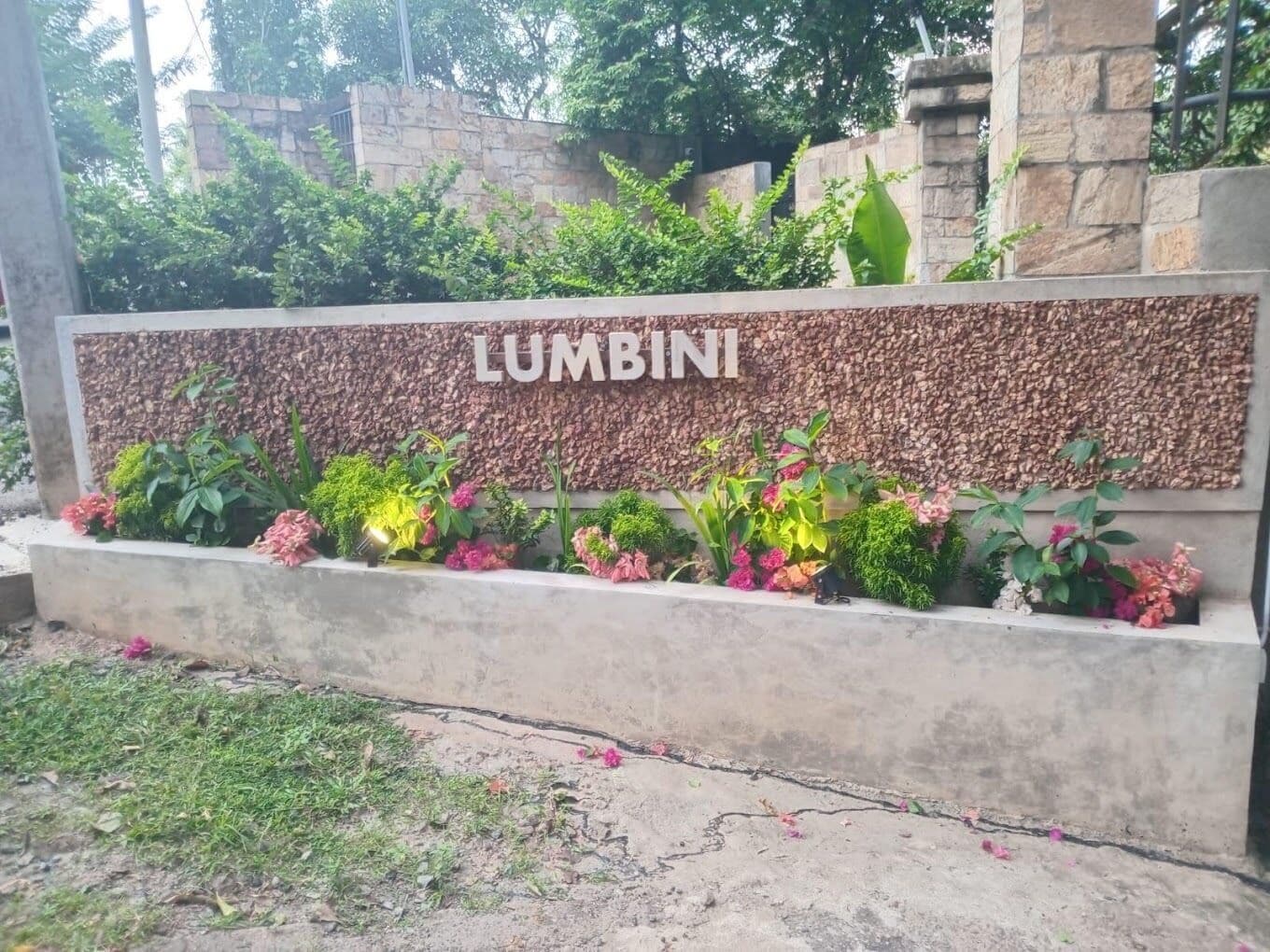 A sign with the word 'LUMBINI' on a stone wall, surrounded by a variety of plants and flowers.