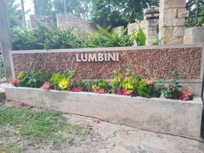 Un letrero con la palabra 'LUMBINI' en una pared de piedra, rodeado de diversas plantas y flores.