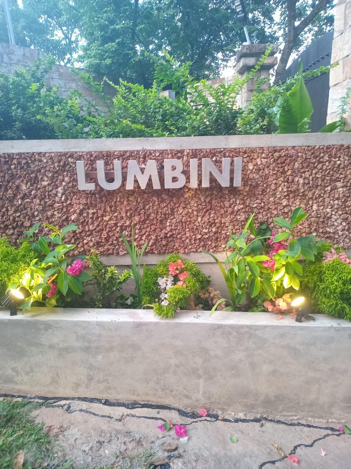 Een bord met de naam 'LUMBINI' op een structuurwand, omgeven door groen en kleine lichtjes.