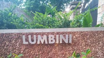 Ein Schild mit der Aufschrift 'LUMBINI' an einer strukturierten Wand, umgeben von Grünpflanzen und kleinen Lichtern.