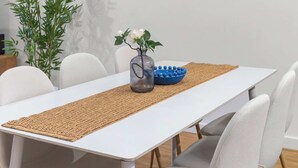 Een eethoek met een witte tafel, grijze stoelen, een geweven onderzetter, een blauwe kom en een wandklok.