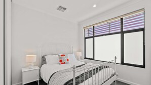 A bedroom with a bed, two bedside tables, a window with blinds, and a ceiling vent. - Campbelltown modern home Family Stay (Campbelltown)