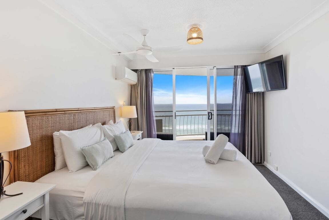A hotel room with a large bed, a ceiling fan, a flat-screen TV, and a balcony with a view of the ocean.