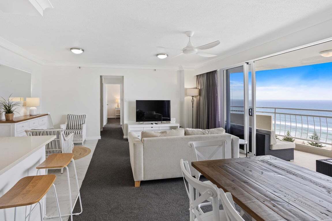 A modern hotel room with a balcony, a flat-screen TV, a sofa, a dining table, and a ceiling fan.