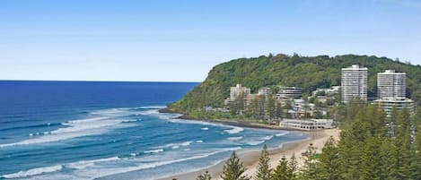 A coastal view with a beach, ocean, and a hill with buildings.
