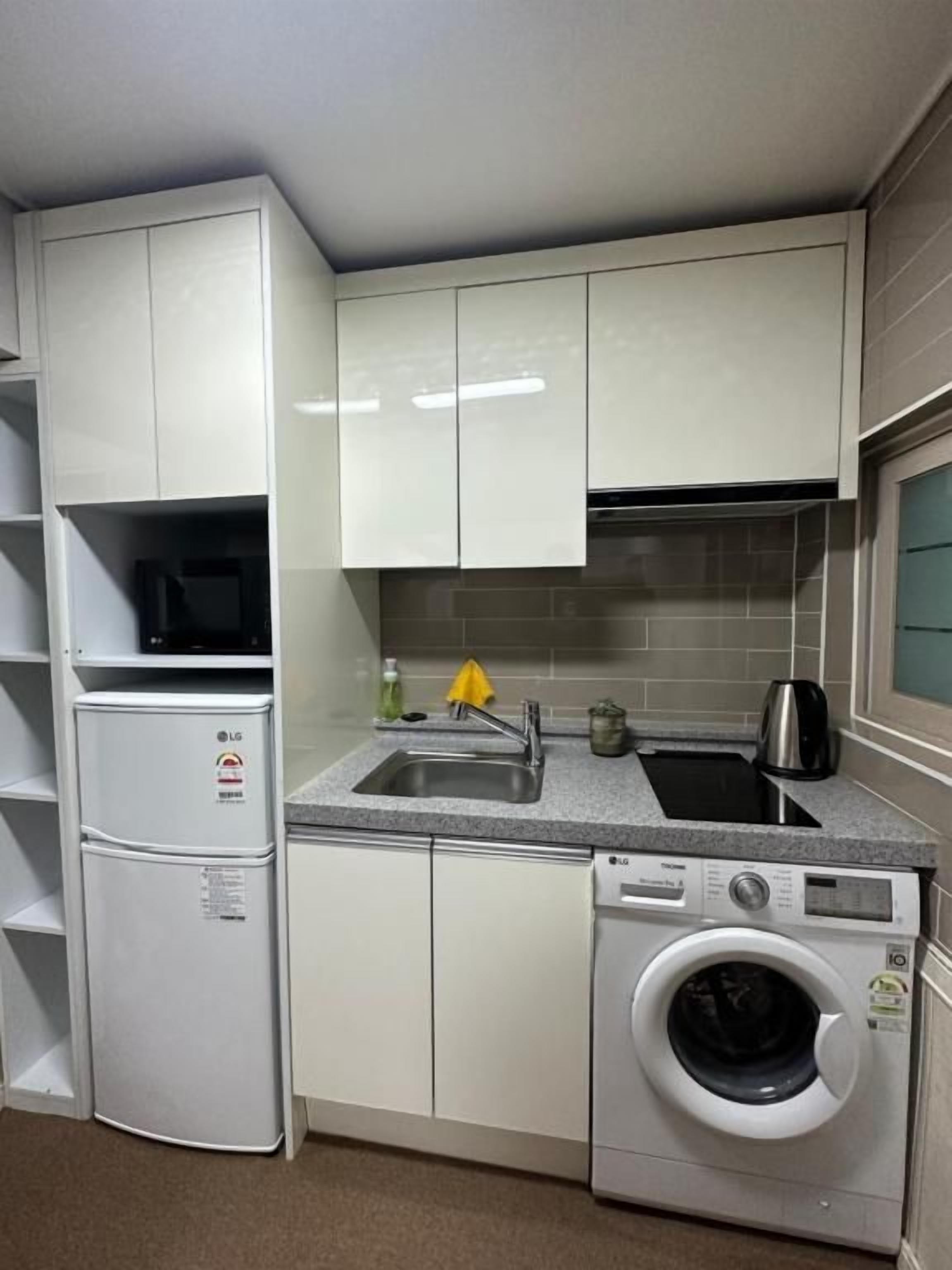 A compact kitchen with white cabinets, a built-in washing machine, and a sink with a kettle.