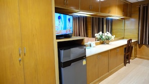 Cocina compacta con armarios de madera, refrigerador, televisor y un pequeño comedor con una silla y un jarrón de flores.