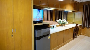 Cocina compacta con muebles de madera, un refrigerador, un televisor y un pequeño comedor con una silla y un florero.