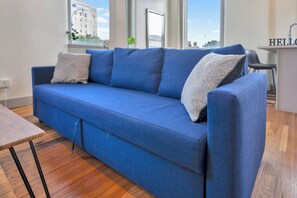 A blue sofa with a textured throw pillow, a wooden table, and a view of a building through a window. - Modern City Loft - Nearby Parking - Downtown - 2F (Louisville)