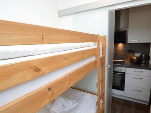 Een stapelbed met houten frame en wit beddengoed, een keuken met witte kasten en een fornuis, en een deur die naar een donkere kamer leidt.