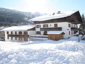 A two-story chalet with a snow-covered roof, surrounded by a snowy landscape and forested hills. - Cozy apartment, opposite the ski lift. (Hinterthal - Maria-alm)