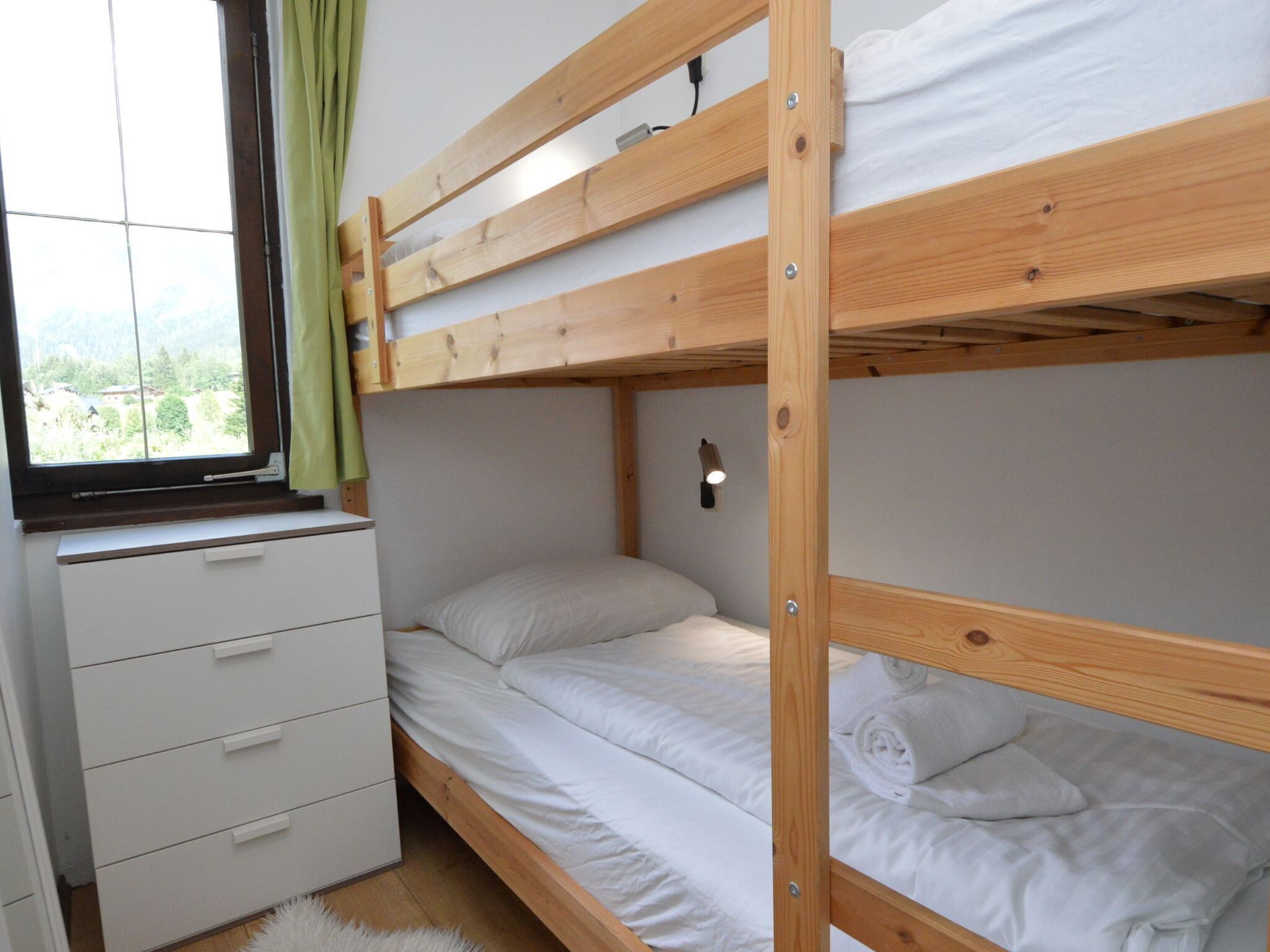 Ein Stockbett mit weißer Bettw äsche, eine weiße Kommode und ein Fenster mit Blick auf Grünflächen.