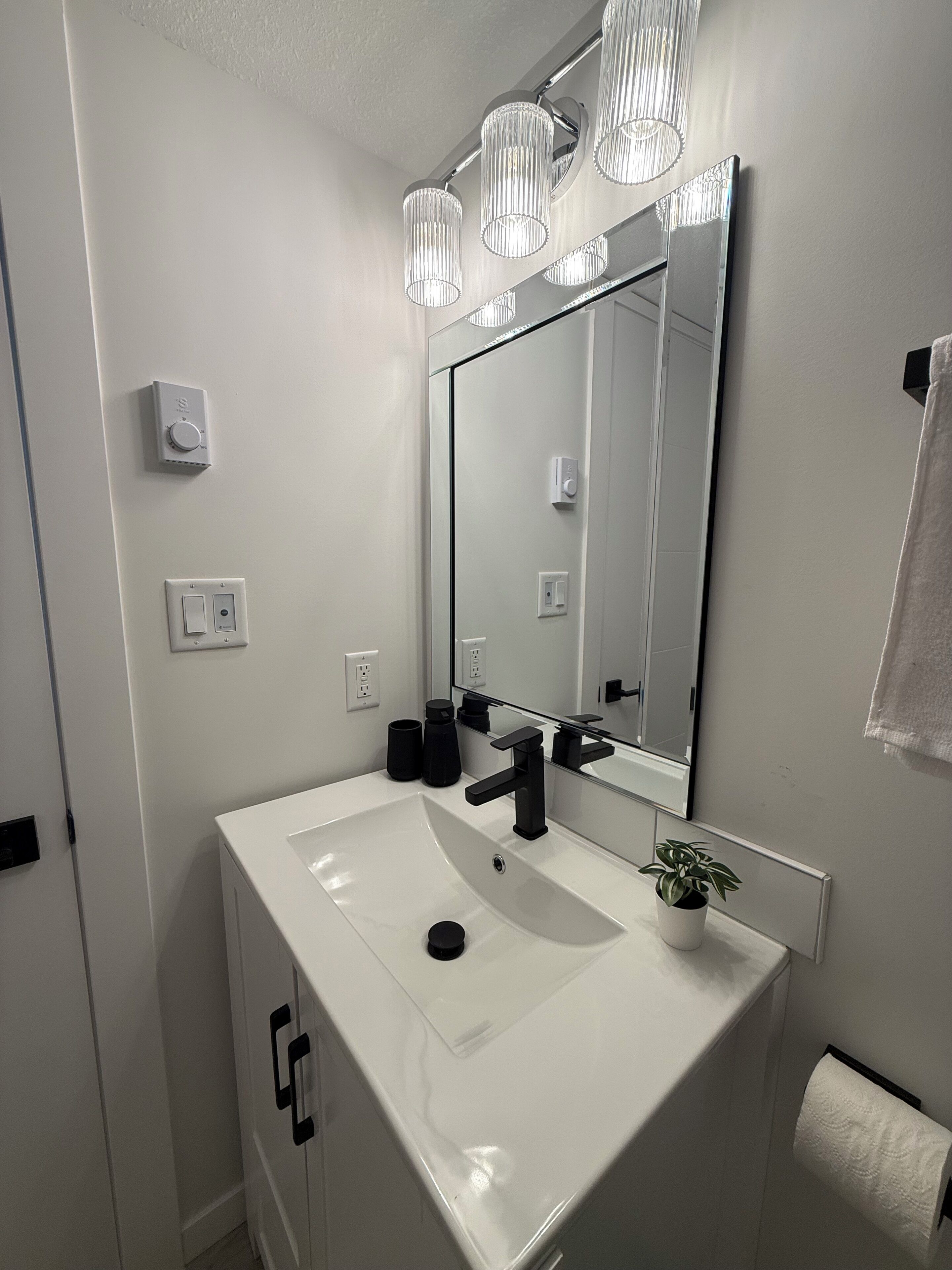 A bathroom with a white sink, black faucet, and a mirror above it. There are three light fixtures above the mirror, and a towel hanging on the wall.