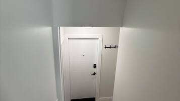 Un pasillo estrecho y bien iluminado con una puerta al final, un felpudo negro al pie y una pared blanca en un lateral.