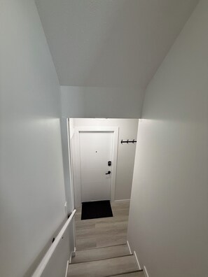 A narrow, well-lit hallway with a door at the end, a black mat at the bottom, and a white wall on one side. - Exquisite and modern one bedroom suite (Regina)