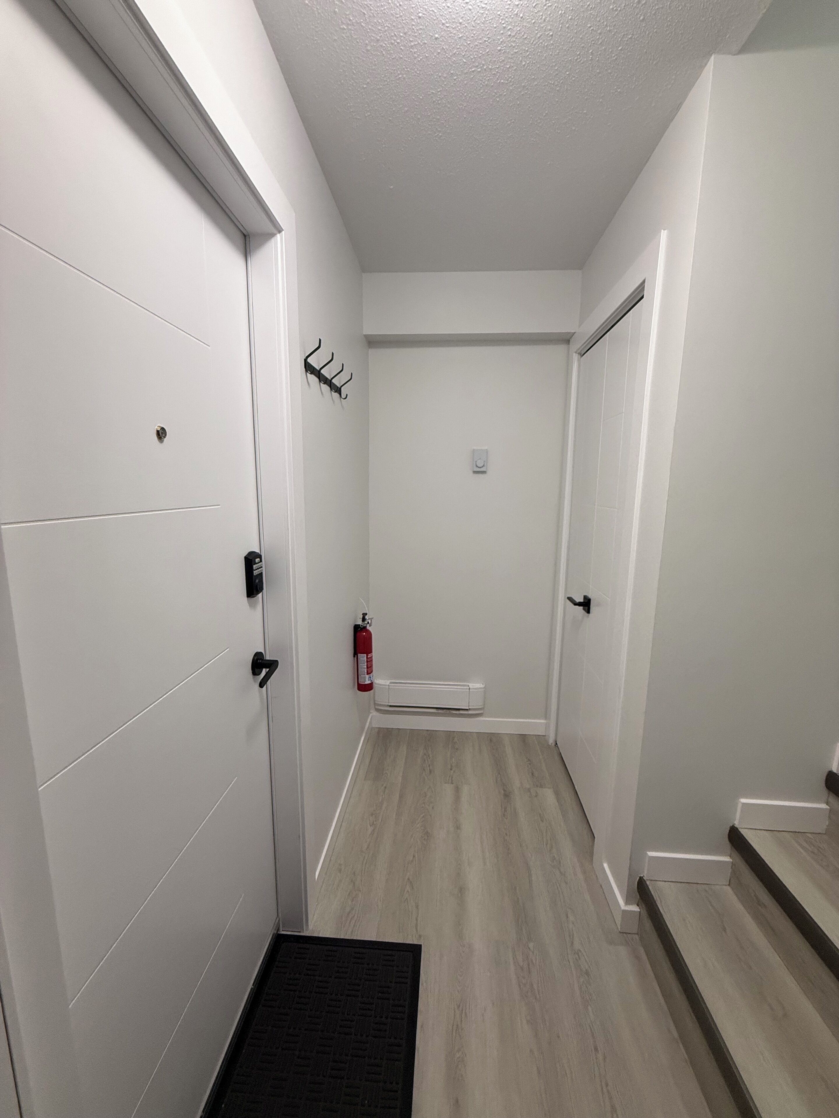 Un pasillo estrecho con paredes blancas, un extintor montado en la pared y una puerta con picaporte negro.
