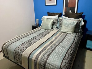 Un lit avec une couvre-lit à motifs et plusieurs oreillers, une table de chevet avec une lampe, et un mur bleu orné d’œuvres d’art.