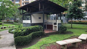 A covered outdoor seating area with a grill, surrounded by a well-maintained garden and benches. - Jaco, Beautiful apartment just a few steps from the ocean, perfect for families. (Jacó)