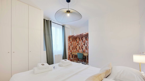 A hotel room with a bed, a desk, and a chair. There is a window with curtains, a wall with a leaf pattern, and a ceiling light fixture. - Cosy Appartement - 1bdr/4p - Boulogne Billancourt (Boulogne-Billancourt)