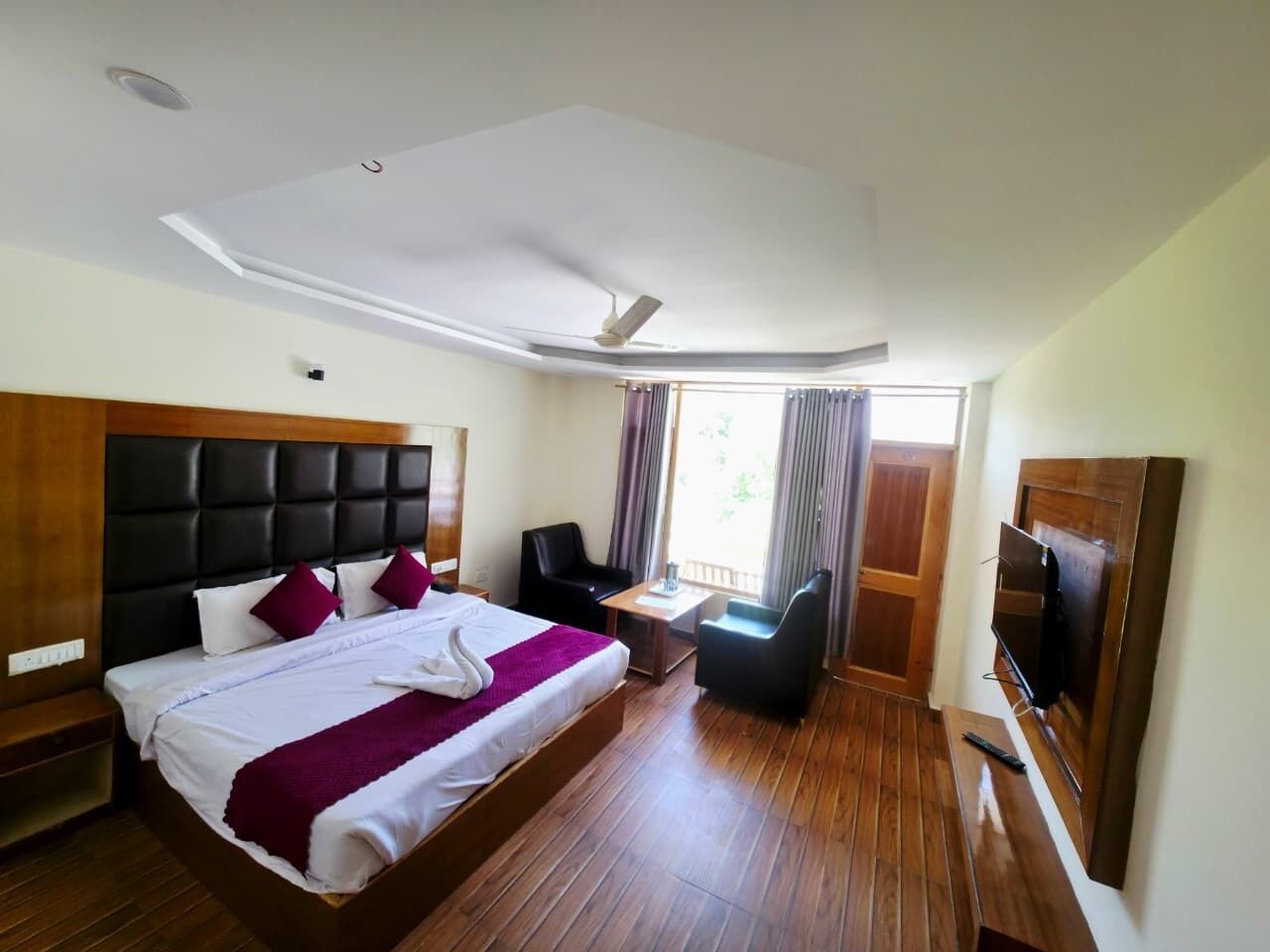 A hotel room with a large bed, a desk, a chair, a TV, and a balcony with curtains.
