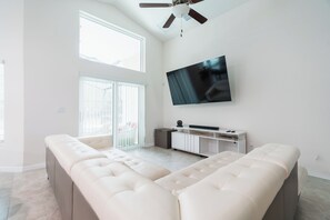 A spacious living room with a large flat-screen TV mounted on the wall, a sliding glass door leading to a balcony, and a white sectional sofa. - Gorgeous, recently remodeled 4-bedroom cozy home with pool near Disney (Kissimmee)
