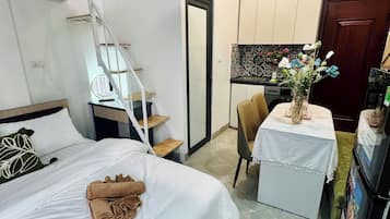 Habitación de hotel con cama, escritorio con silla, pequeña mesa con un florero, refrigerador y proyector en el techo.