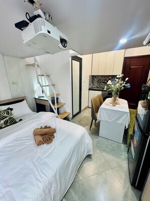 A hotel room with a bed, a desk with a chair, a small table with a vase of flowers, a refrigerator, and a ceiling projector.