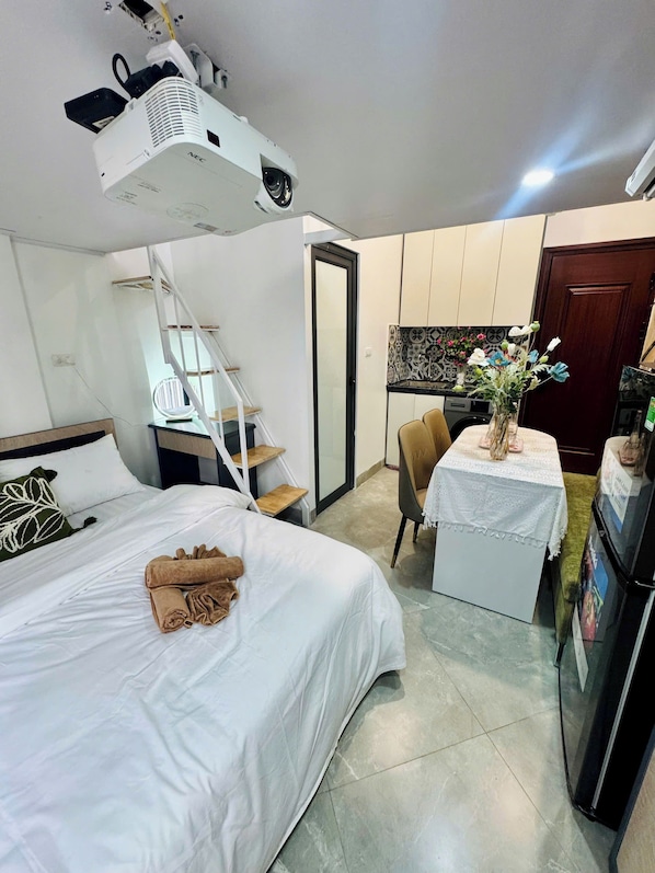 A hotel room with a bed, a desk with a chair, a small table with a vase of flowers, a refrigerator, and a ceiling projector.