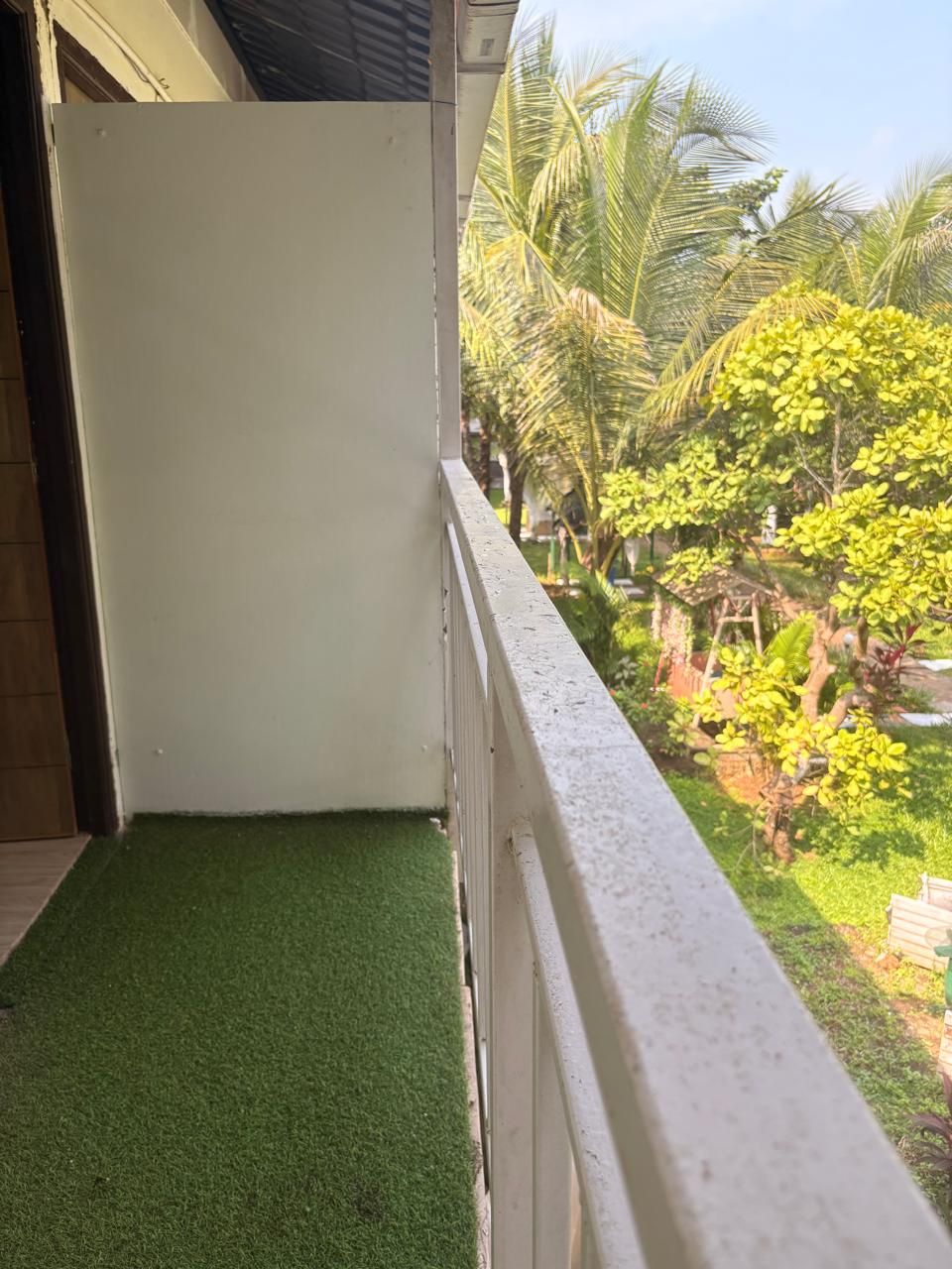 A balcony with a white railing, a green artificial grass floor, and a view of a garden with palm trees and yellow flowers.