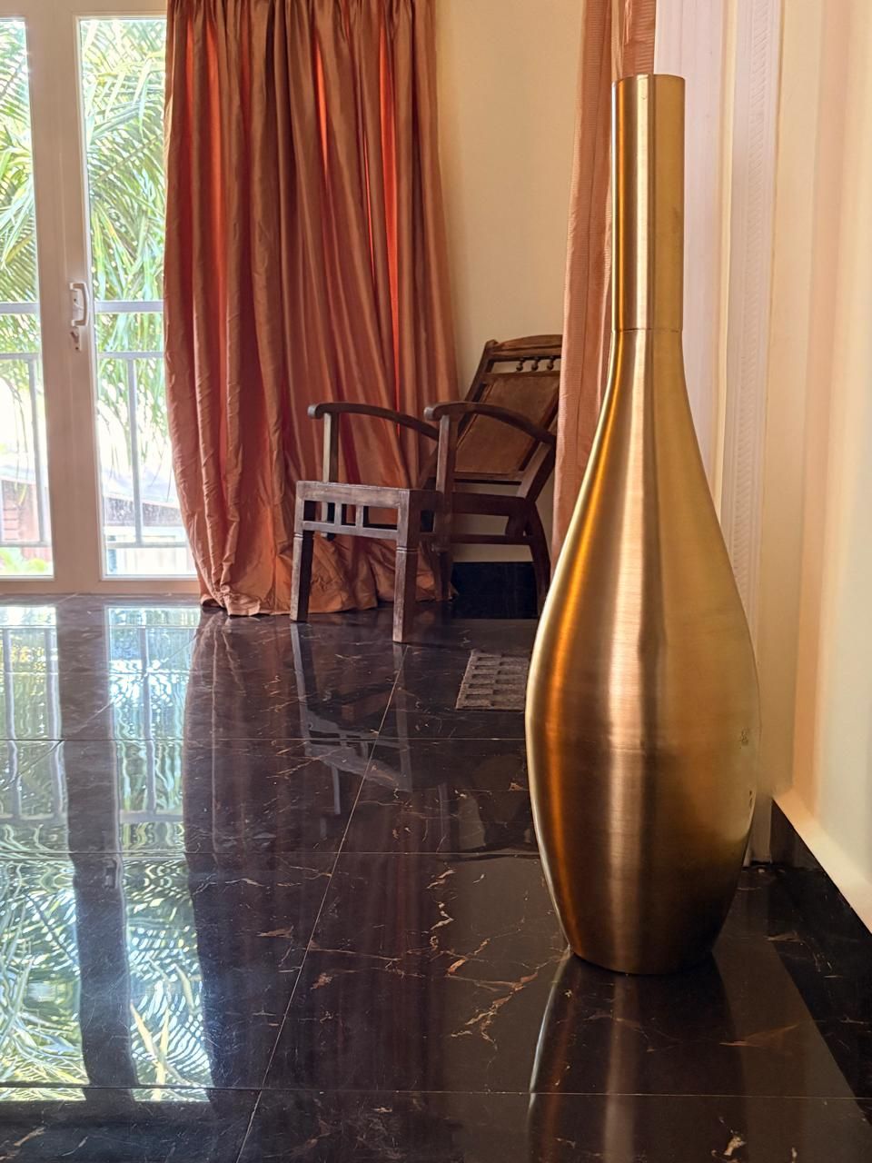 Une pièce dotée d’un sol carrelé noir brillant, d’un grand vase métallique, d’une chaise en bois et d’une fenêtre avec des rideaux.