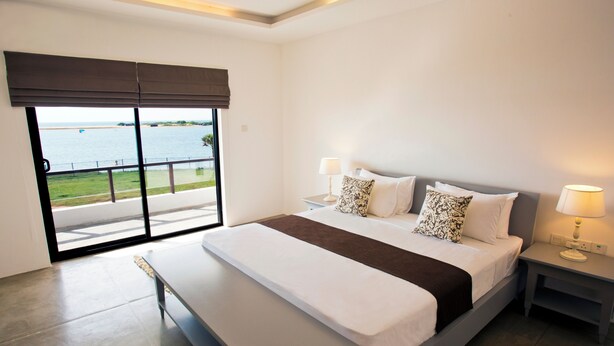 A modern bedroom with a large bed, two bedside tables with lamps, a bench, and a view of a body of water through a sliding door.