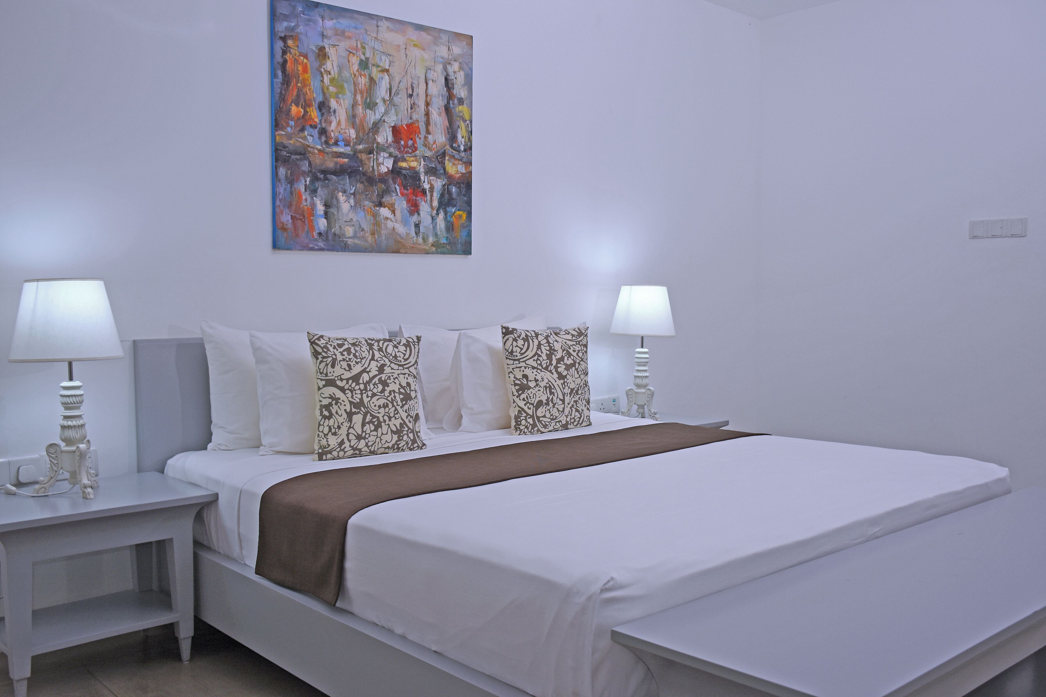 A hotel room with a bed, two bedside tables with lamps, a large abstract painting on the wall, and a brown throw blanket.