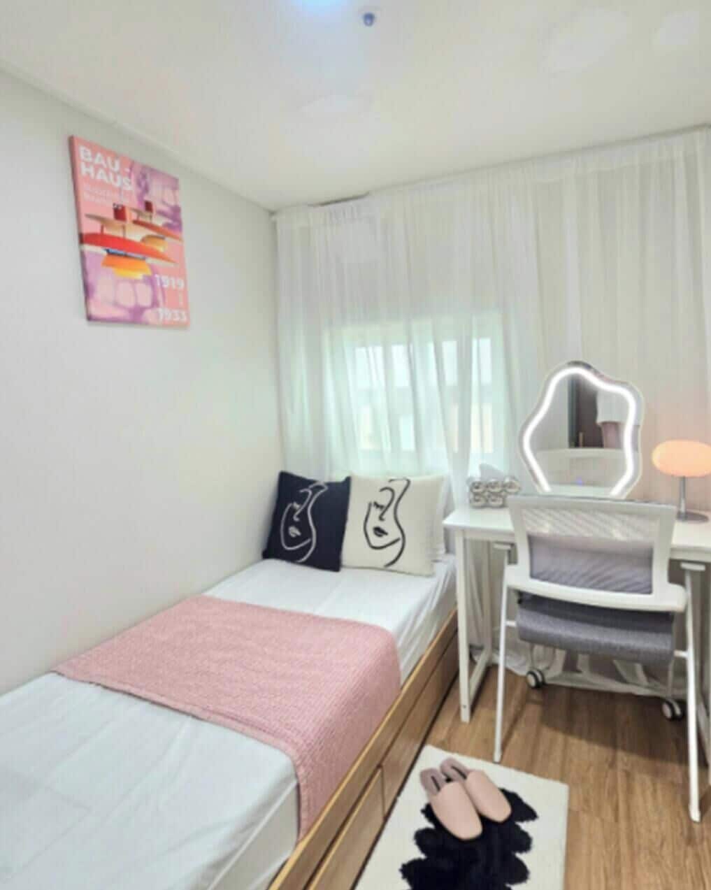 A hotel room with a bed, a desk with a mirror, a chair, and a pair of slippers on the floor.
